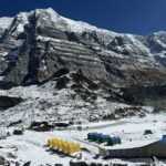 Campamento de tiendas sobre morrena con cumbres nevadas iluminadas al fondo